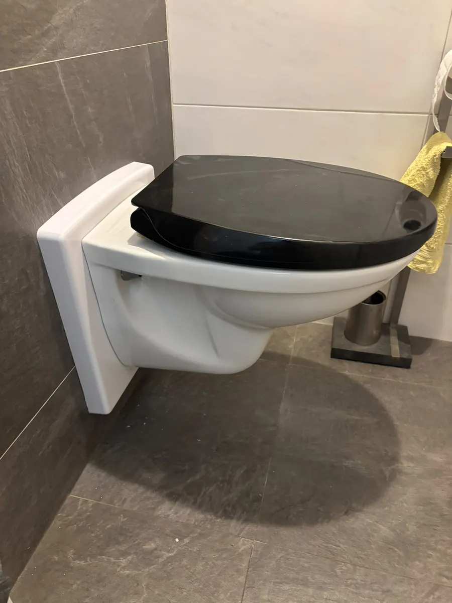 Hangtoilet met zwarte zitting op een wit verhogingsframe tegen een grijze tegelwand, verhoogd door Van Wouden Installatietechniek